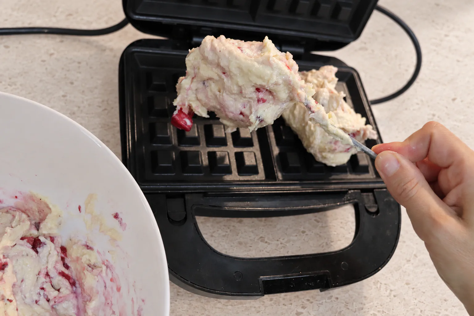 Berry-speckled waffle batter being spooned into a heated waffle maker